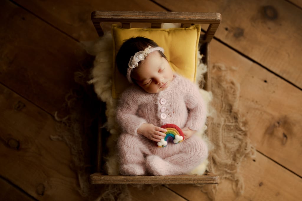 newborn baby girl on miniature bed with yellow pillow holding rainbow prop
