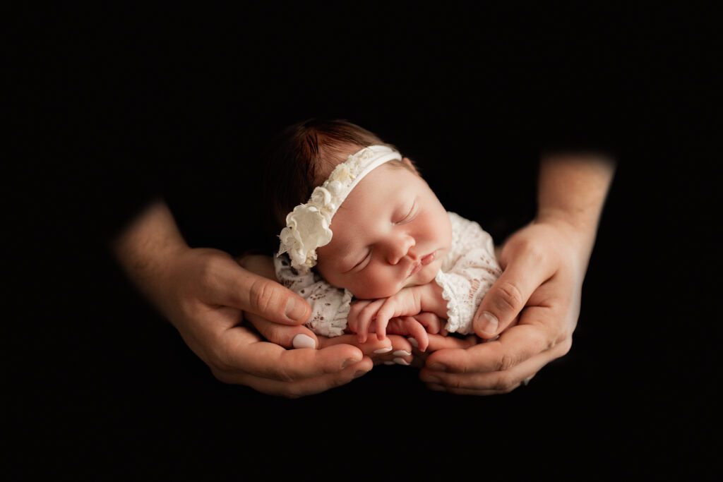 Baby girl in white against black backdrop, cradled in parents hands