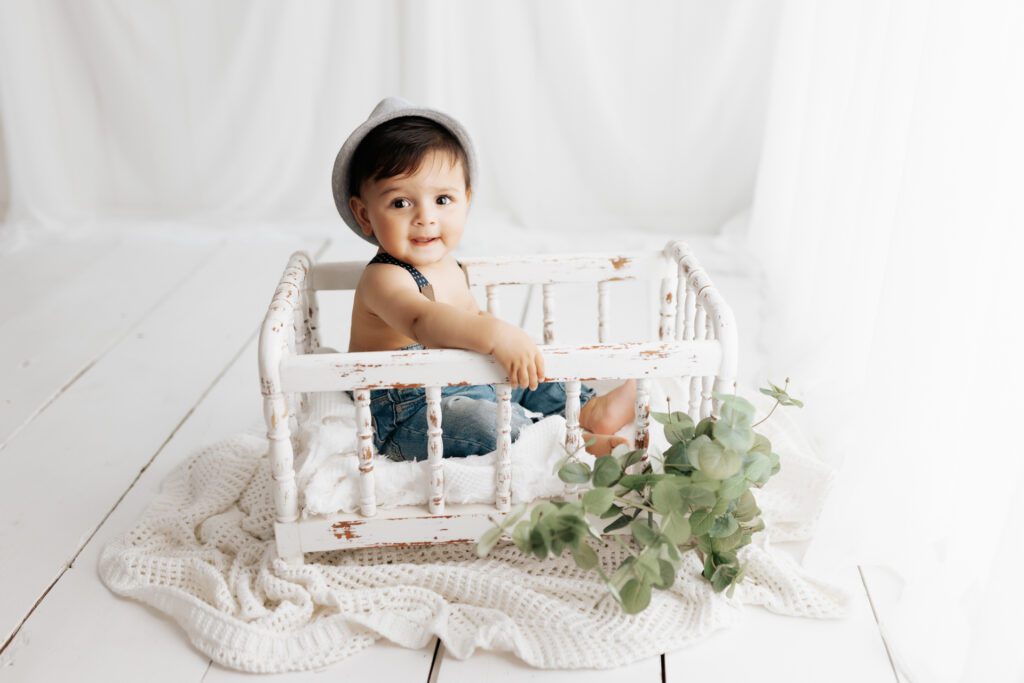 Baby boy in overalls and hat seated in miniature crib in Long Grove, Illinois photo studio