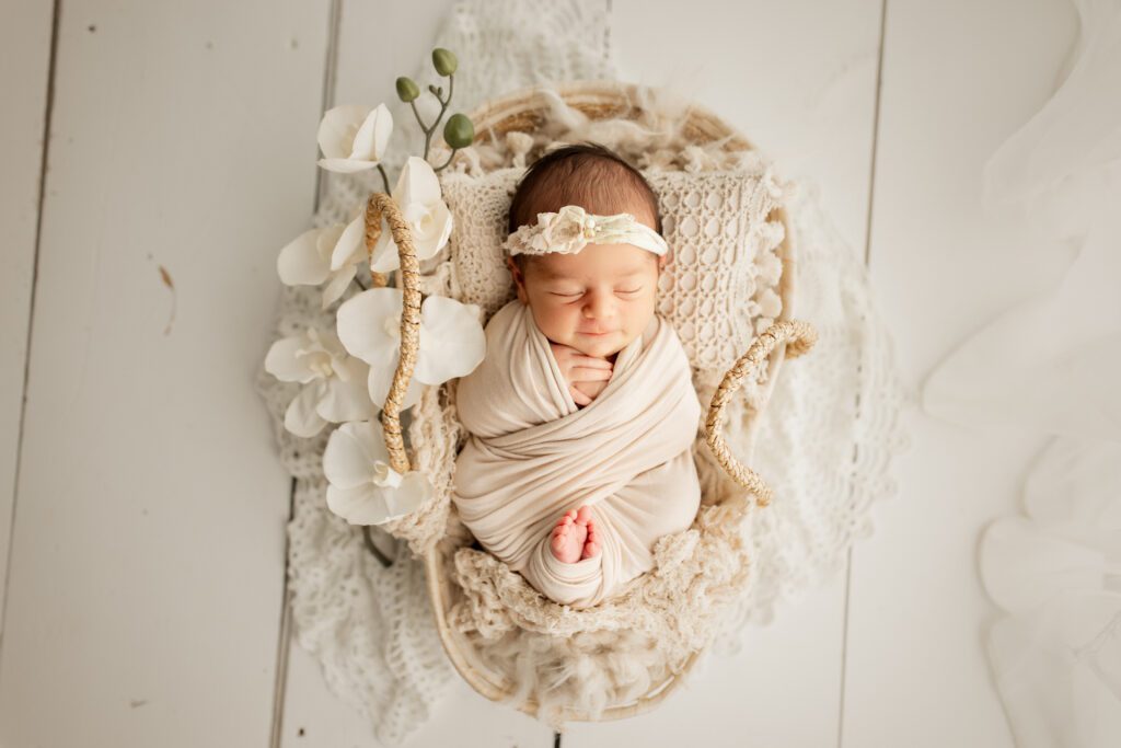 Baby girl in cream wrap asleep in basket with white flowers next to her