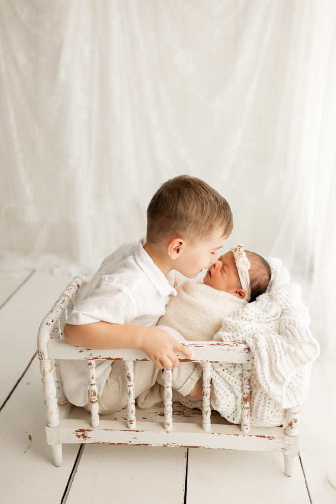 Boy seated in distressed white crib giving new baby sister a nose kiss