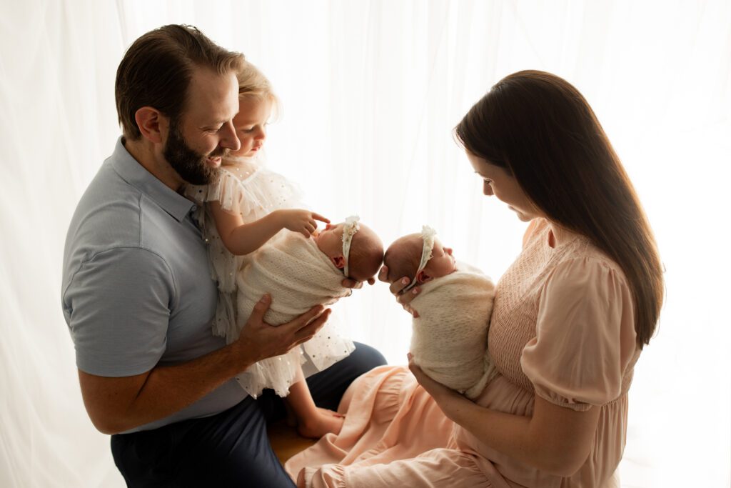 Parents and toddler holding newborn twin girls near window in Long Grove Illinois photo studio