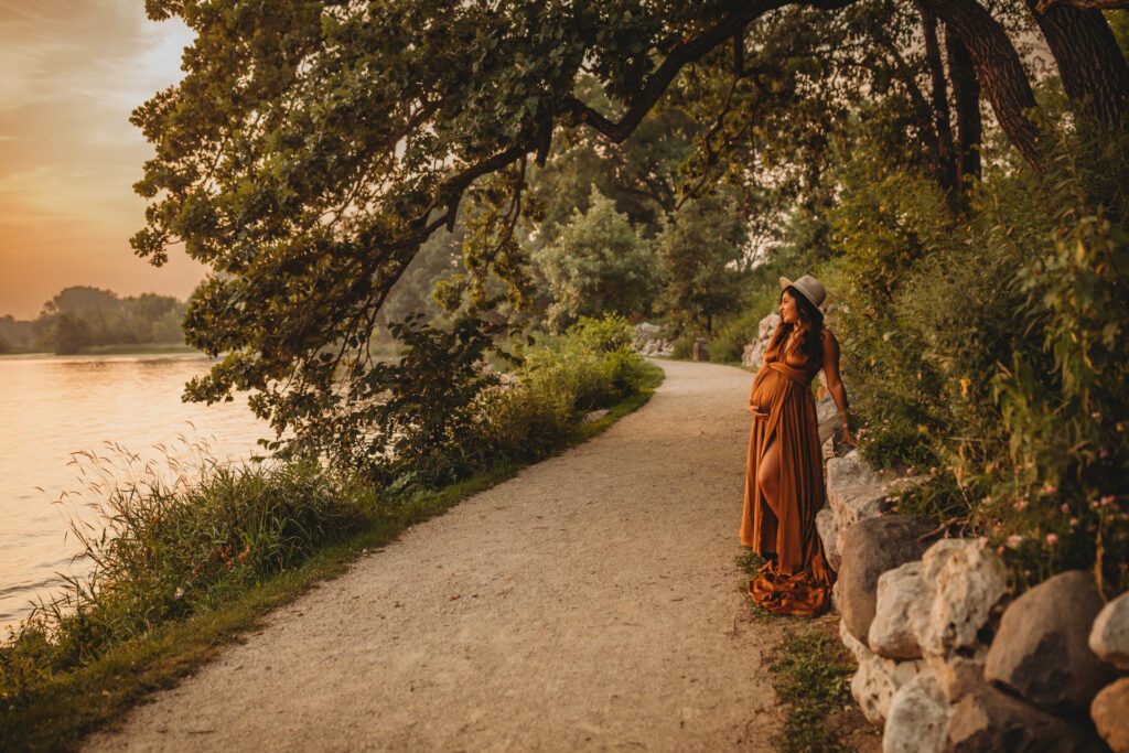 Woman in rust-colored maternity gown looking across a lake at sunset