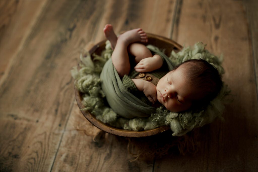 Newborn baby boy swaddled in green and lying asleep in a wooden bowl