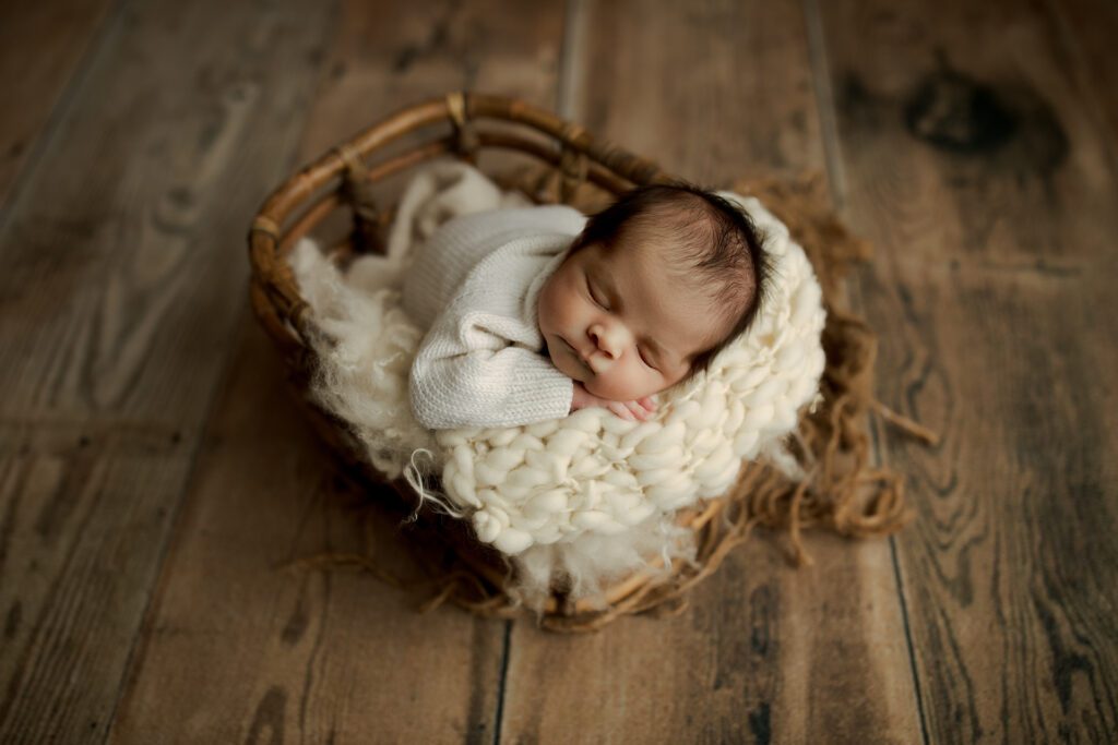 Newborn boy asleep in basket against wooden flooring backdrop in Chicagoland photo studio