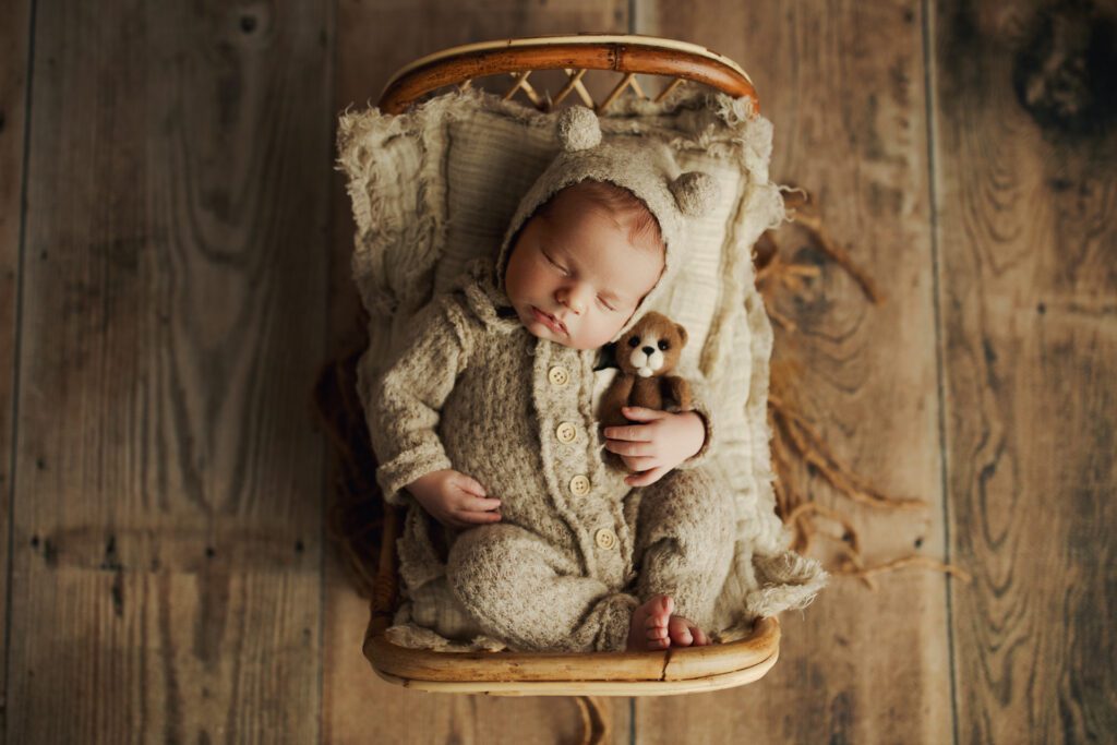 Newborn boy in bear cap and pajamas asleep with teddy bear in miniature bed