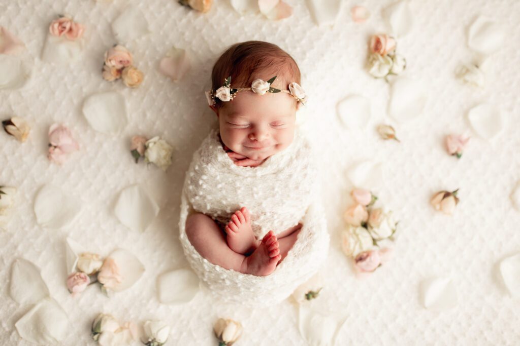Smiling newborn girl asleep in white swaddle and surrounded by rose petals