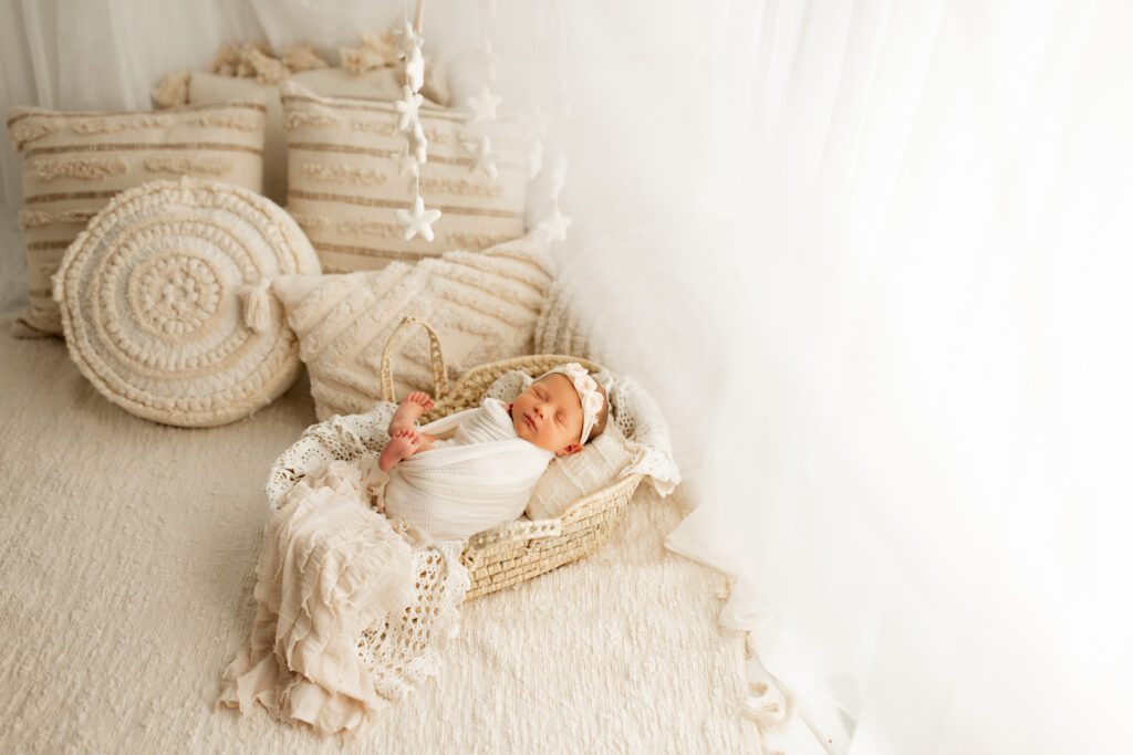 Swaddled newborn girl in basket lying next to cushions with star mobile hanging above her