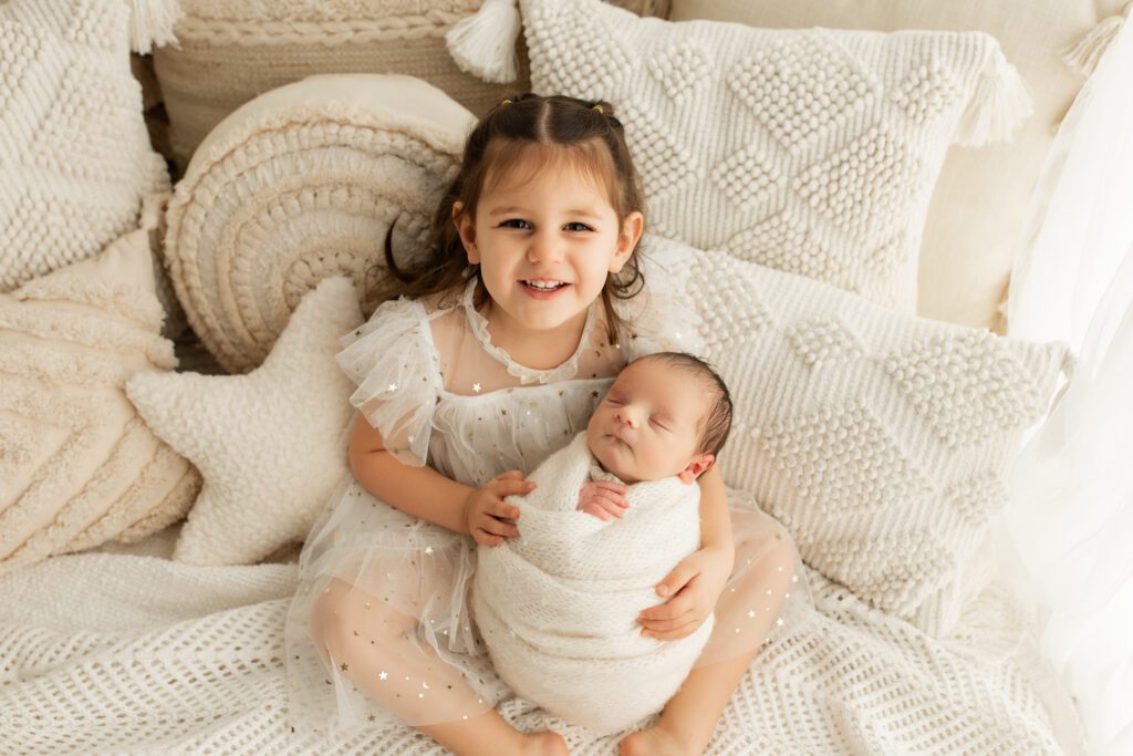 Little girl in white dress seated on bed in Chicagoland photo studio holding new baby brother