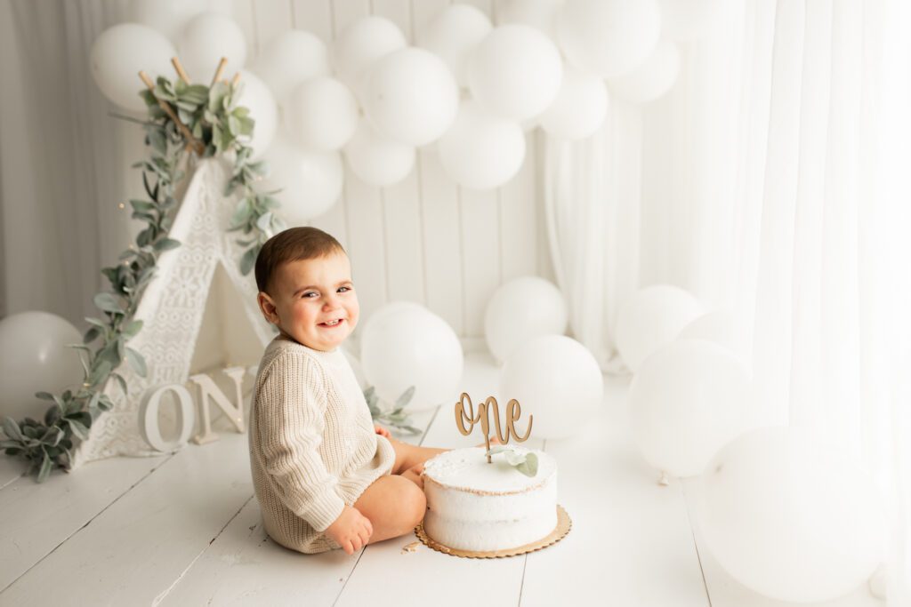 Baby in onesie seated next to cake and smiling at camera in studio near Chicago