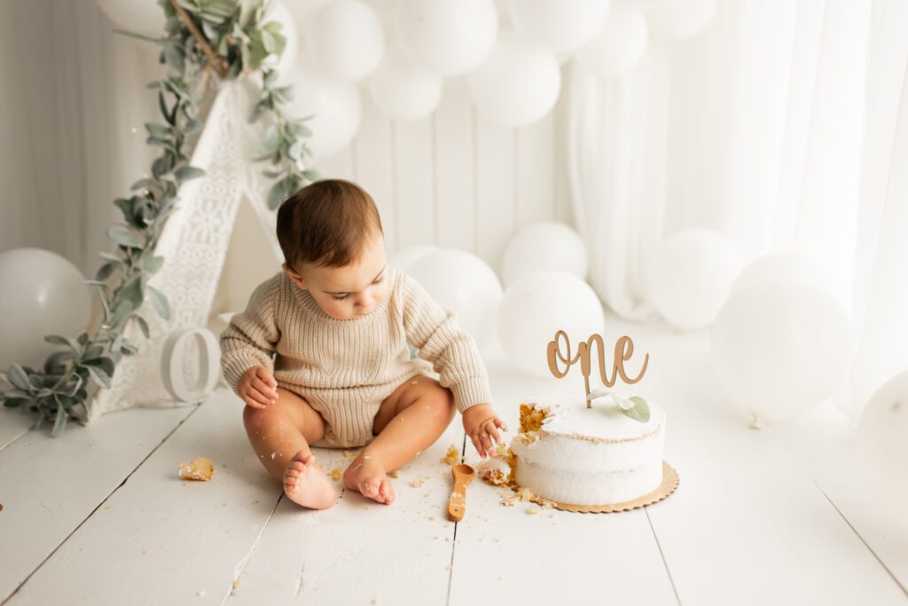 One-year-old baby exploring cake with topper against background of white balloons
