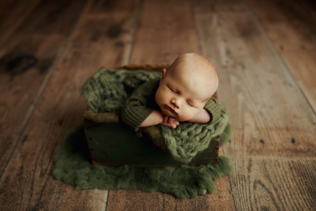 Infant boy asleep in wooden box cushioned with green blankets during Park Ridge newborn photography session