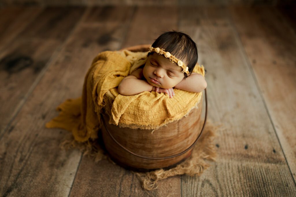 Baby girl in yellow wrap asleep in antique bucket during Chicagoland newborn photoshoot