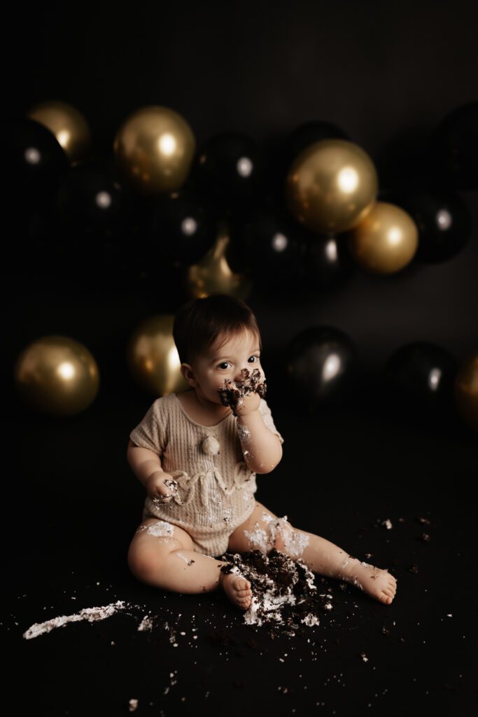 Baby boy eating chocolate cake against backdrop of gold balloons in Long Grove photo studio