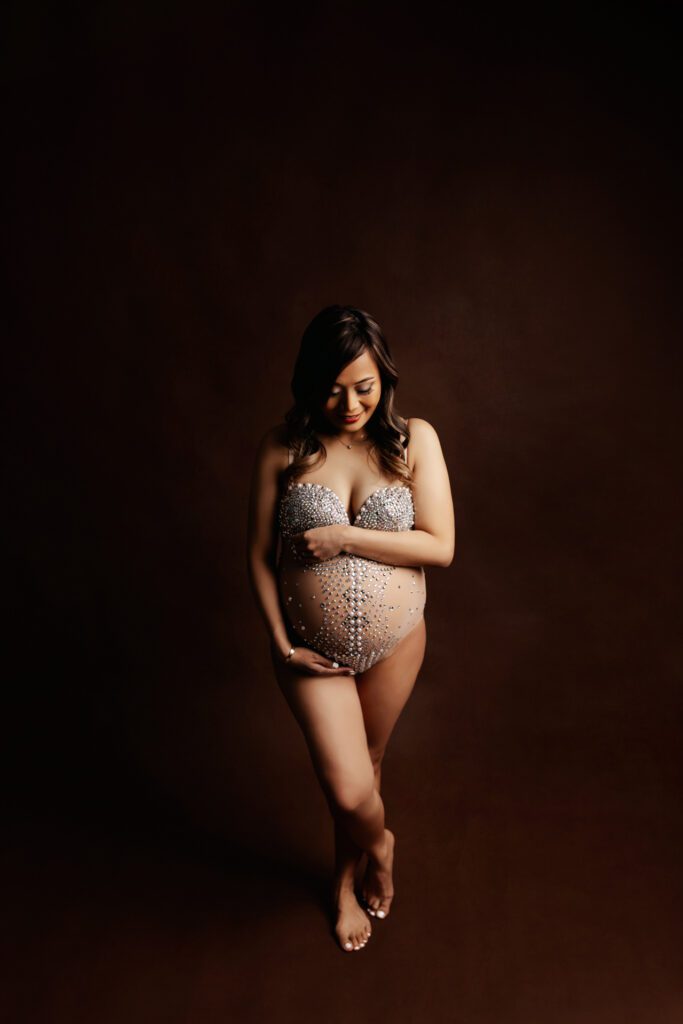Woman in silver sequined maternity bodysuit poses with crossed legs in Chicago photo studio