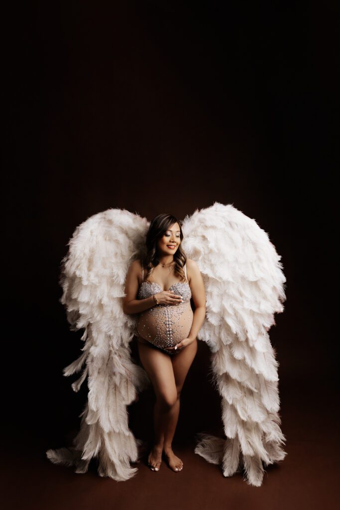 Woman in silver sequined bodysuit and white angel wings cradles her pregnant belly