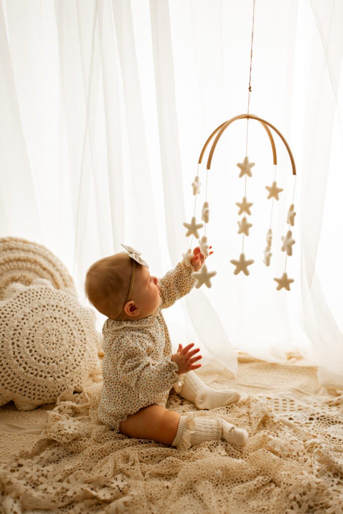 Baby girl in floral print onesie reaches up to touch a star mobile in photography studio near Mount Prospect Illinois