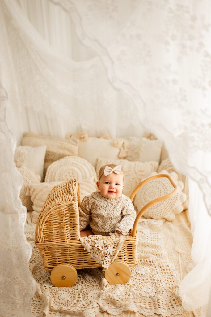 Baby girl in wheeled wicker basket in idyllic nursery setup with textured pillows piled high behind her