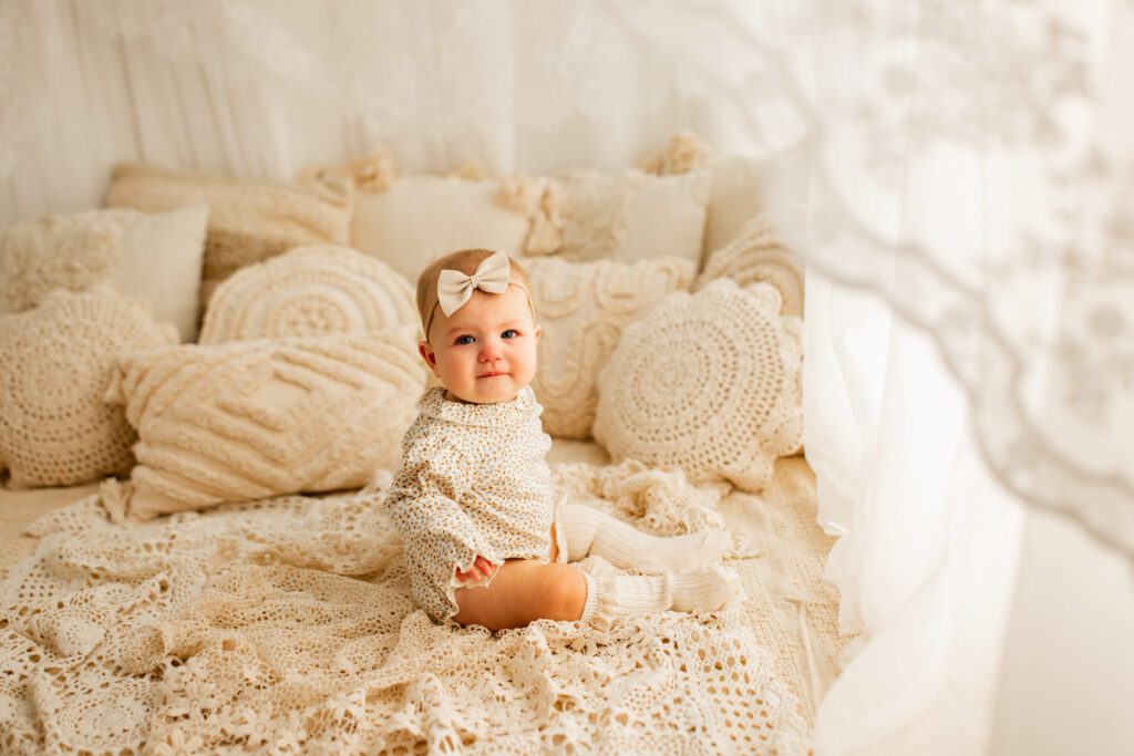Baby girl seated on lacey blanket near curtained window with piles of comfy pillows in the background