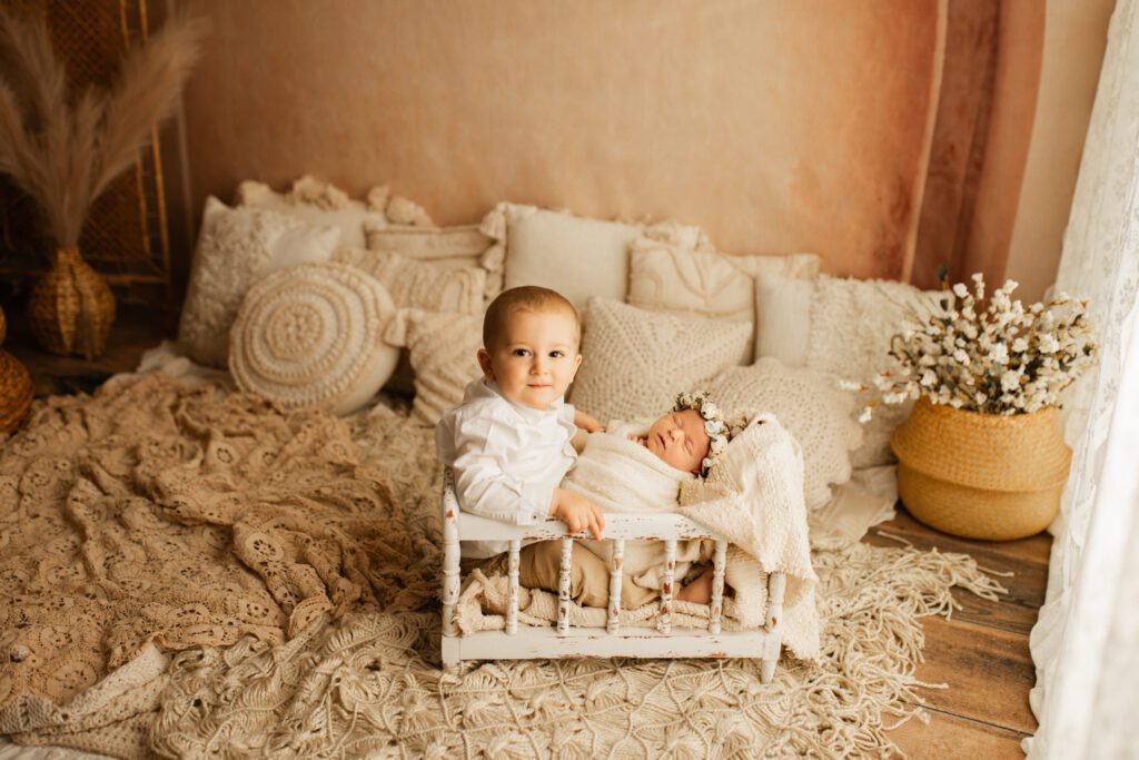 Toddler boy holds his new baby sister on his lap in a miniature crib