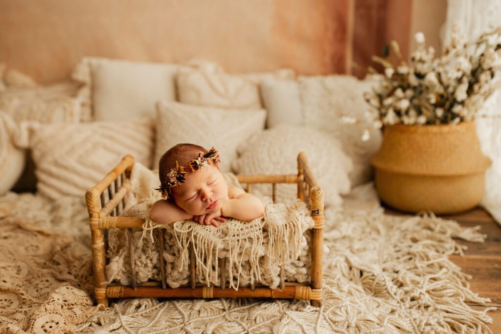 Baby girl asleep in mini crib surrounded by pillows and rugs, with a potted plant in the background