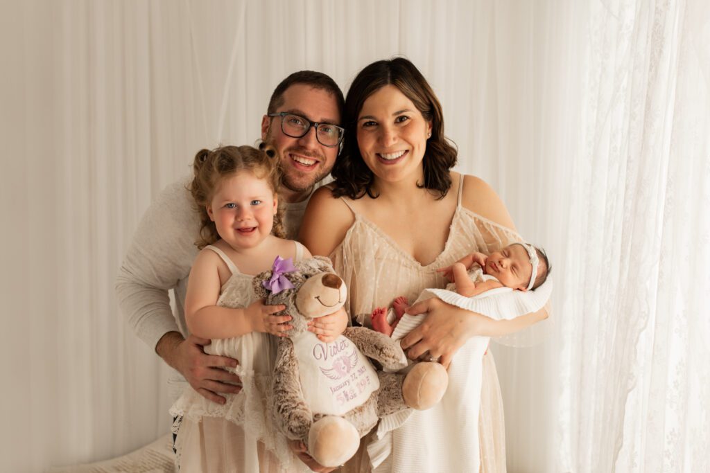 Parents stand with their toddler and newborn daughters near a window with white lace curtains