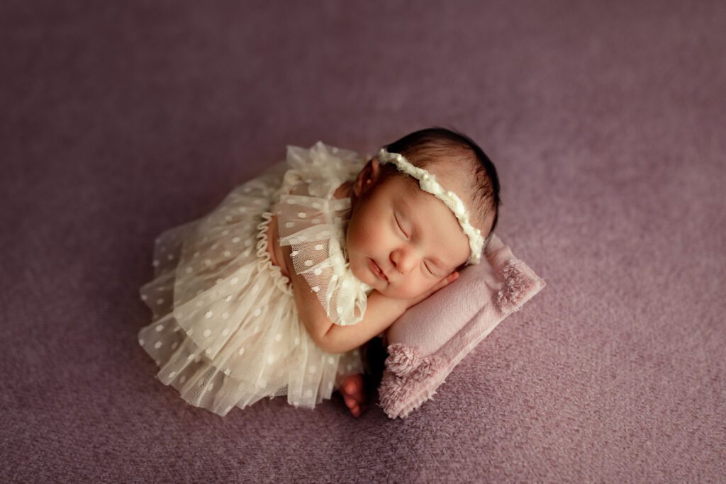 Newborn girl in white polka dot tulle dress curled up asleep on a lavender pillow