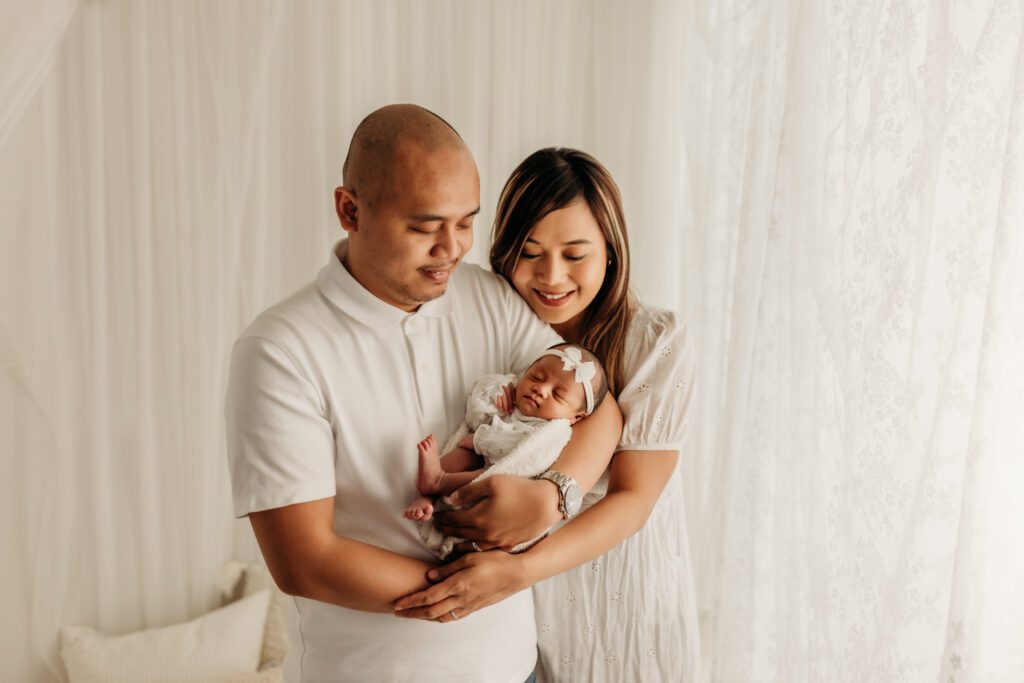 Woman wraps her arms around her husband as they both gaze down at the baby girl asleep in his arms