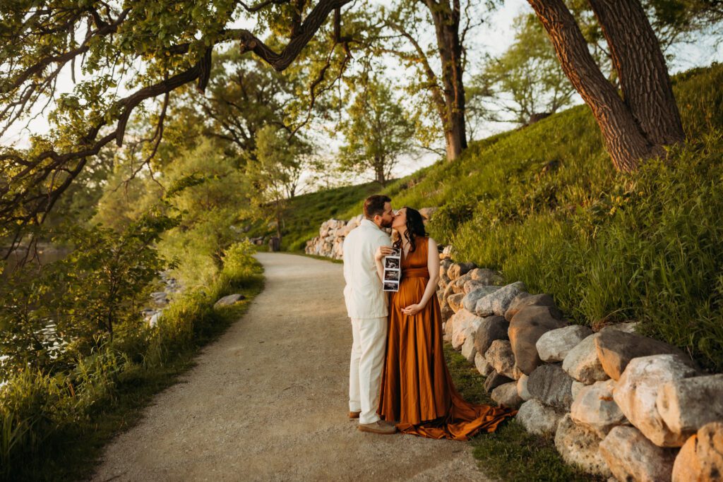 Woman standing on a grassy path holding sonogram pictures of her baby while she and her husband exchange a kiss