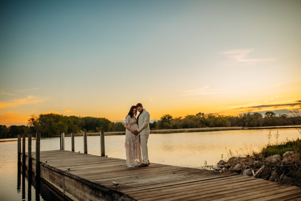 Couple hold hands on a wooden lake dock at sunset during a maternity session with one of Chicago's best maternity photographers