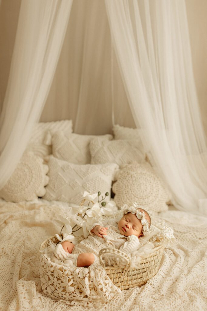 Baby girl wearing flower crown tucked up in basket surrounded by pillows and lacy blankets