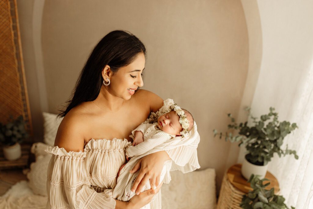 Mother in boho dress stands in a cozy nook by a window cradling her baby in her arms