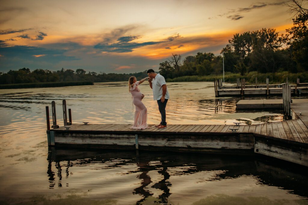 Man kisses his pregnant wife's hand as they stand together on a lakeside dock near Chicago at sunset