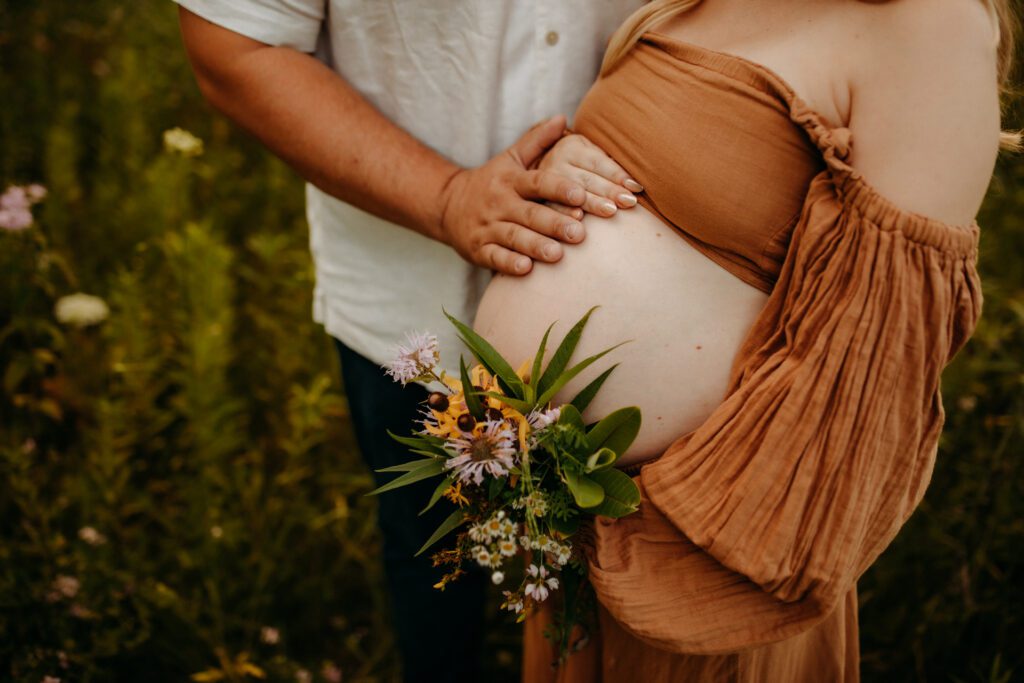 Pregnant woman holding a bouquet of wildflowers as she and her husband lay their hands on her belly