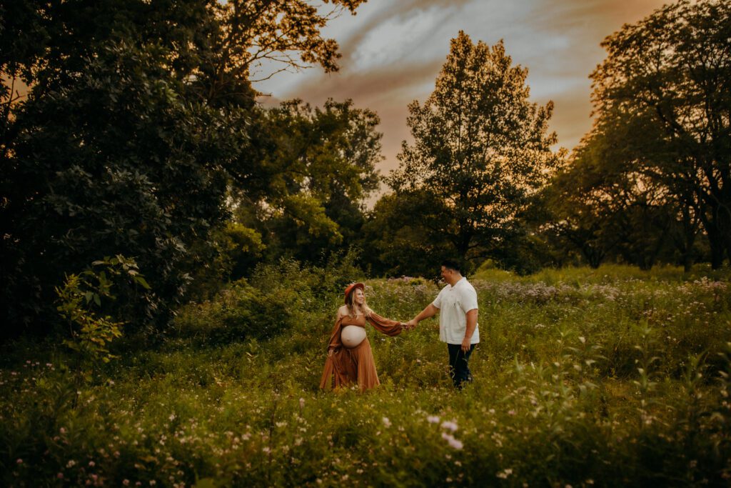 Pregnant woman and her husband walk together through a tree-lined meadow at sunset during their session with one of Chicago's best maternity photographers