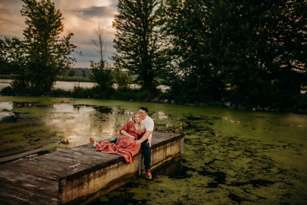 Pregnant woman in boho maternity dress leans back against her husband as they sit together on a dock at the edge of a lake