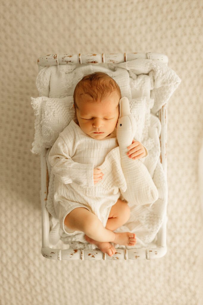 Newborn boy in white linen onesie snuggles with a duck stuffie during his newborn photoshoot near Palatine Illinois