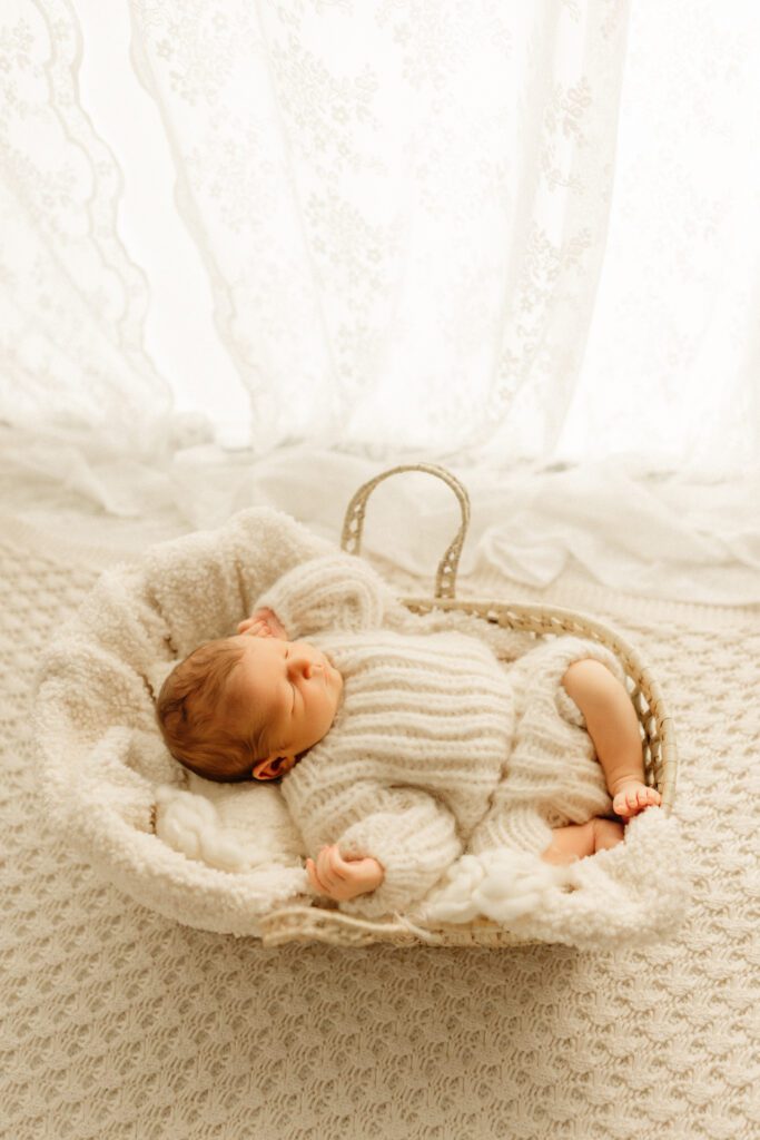 Newborn baby boy in chunky sweater sleeps peacefully in a basket near a curtained window during his newborn photo session