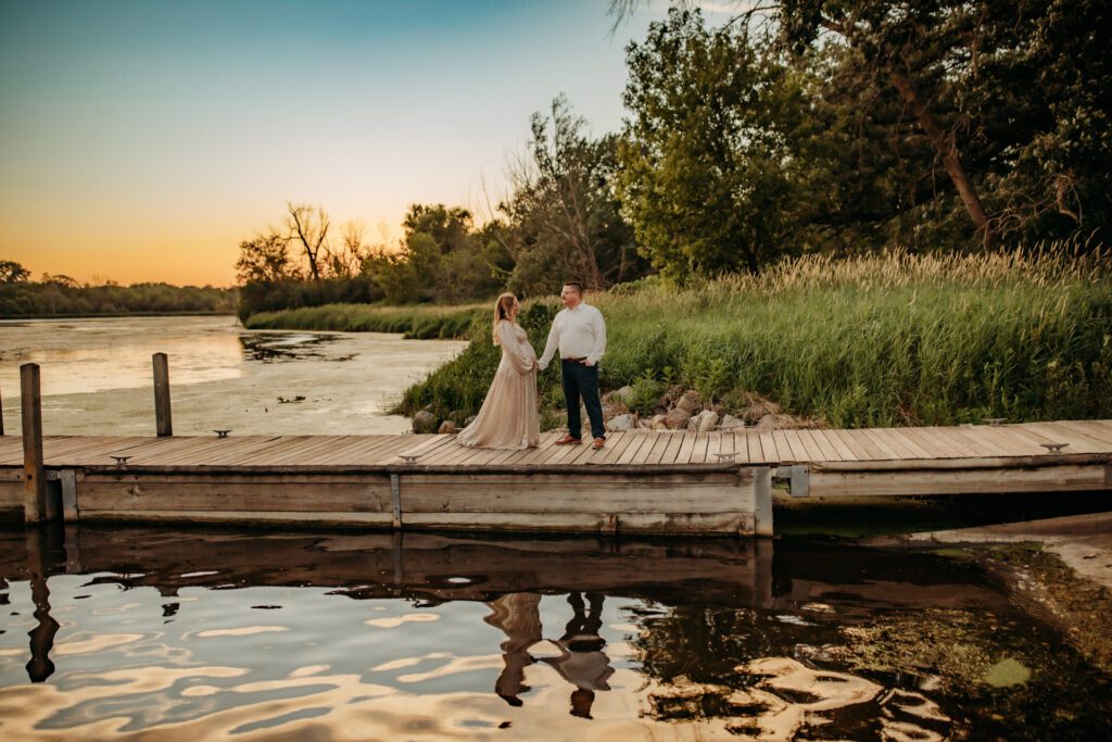 Couple holding hands on a lakeside dock at sunset as they pose for maternity pictures near Chicago