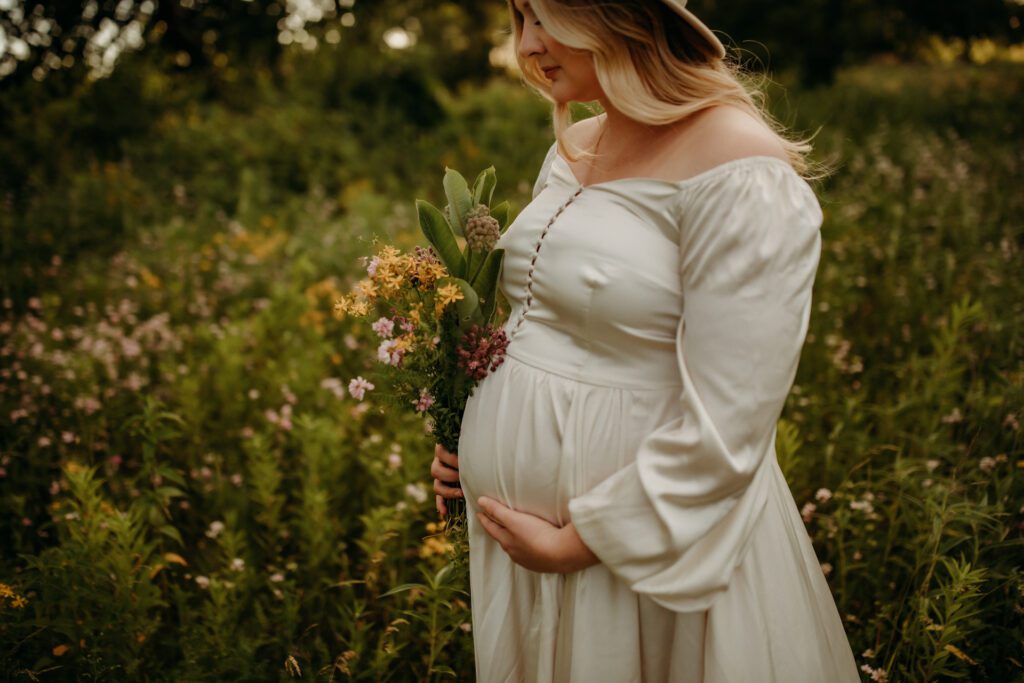 Woman in flowing white dress cradles her pregnant belly with one hand while she holds a bouquet of wildflowers in the other