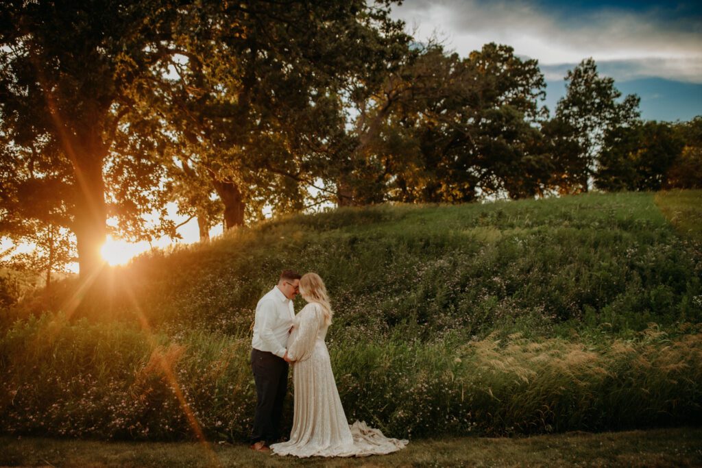 Pregnant woman in beautiful maternity gown touches foreheads with her husband as they stand together in a meadow with the sun setting through the trees