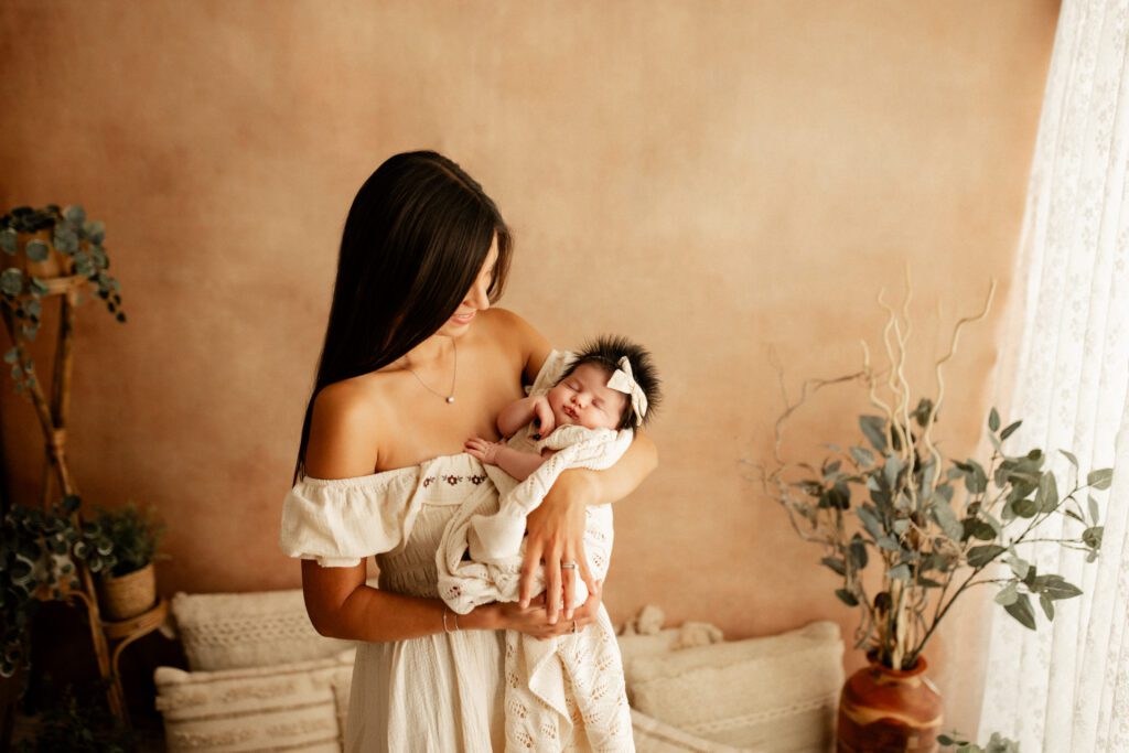 Mother in white dress stands in a curtained alcove decorated with plants, holding her newborn baby girl in her arms