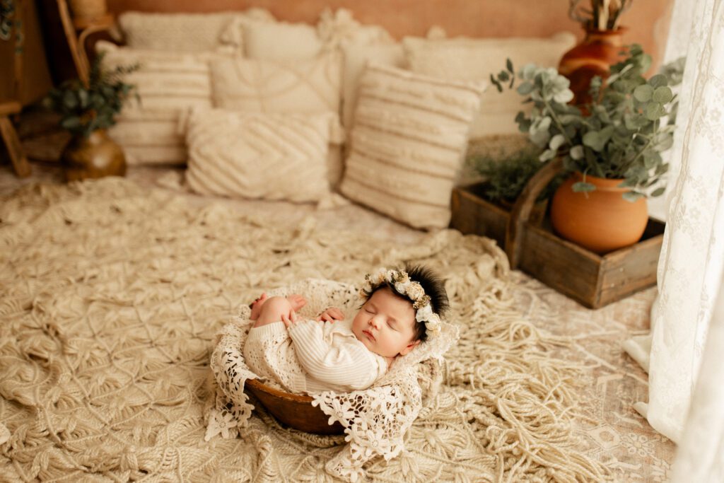 Newborn baby girl with white flower crown asleep in wooden bowl with pillows and plants in the background