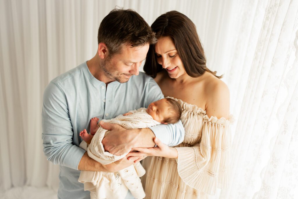 Dad holding his newborn son in his arms while mom embraces them both and they gaze down at baby together