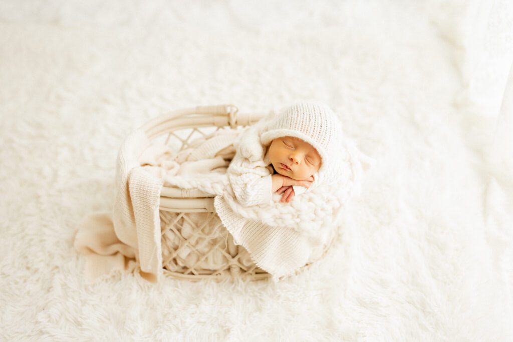 Newborn boy in white outfit and beanie snuggled in a blanket-filled basket during a photoshoot in Chicago studio