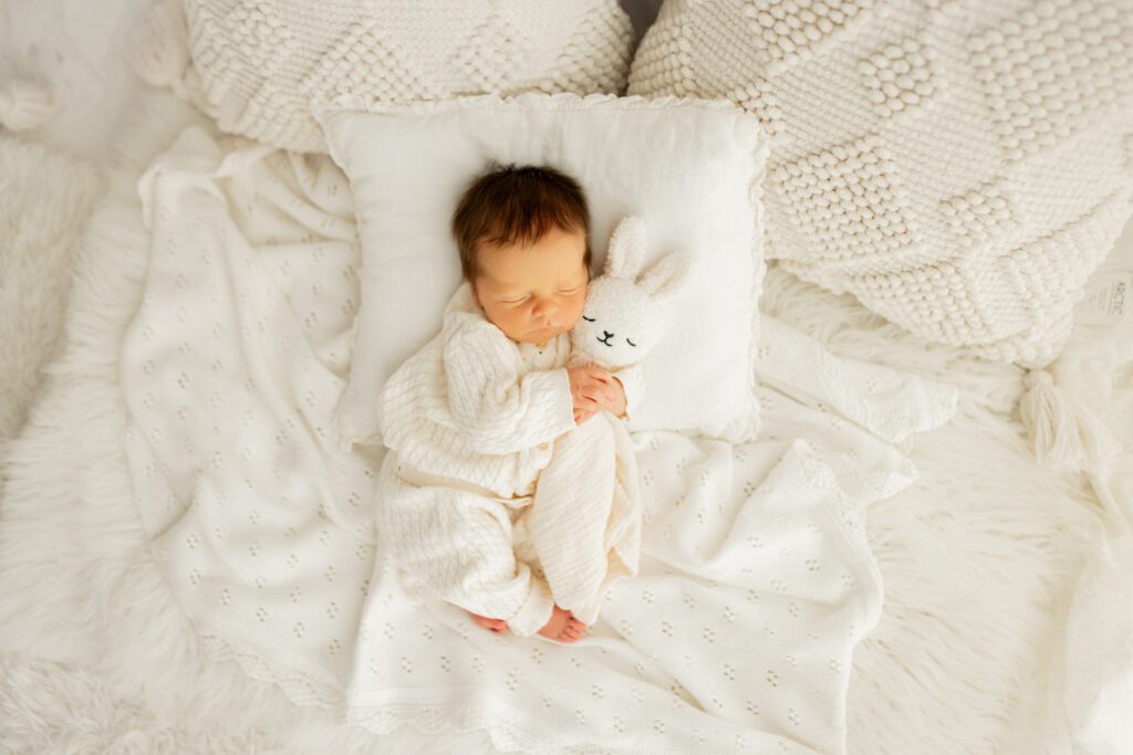Newborn boy in white outfit asleep on a pillow while he cuddles a bunny lovey