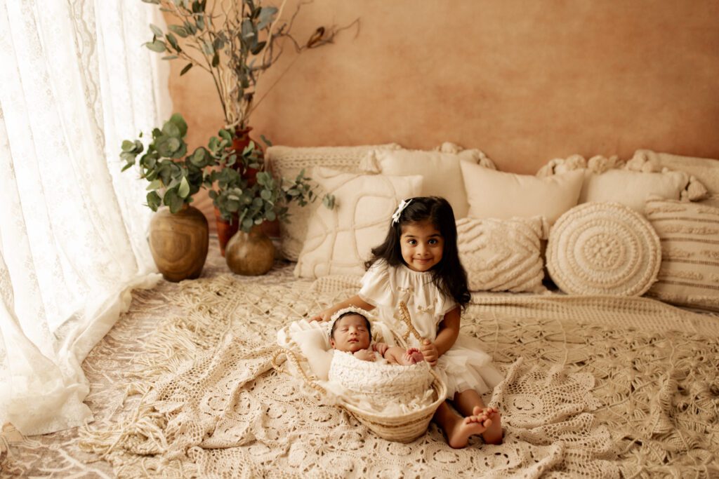 Toddler girl in white dress sits next to her new baby sister in a rustic studio lifestyle setup near Chicago