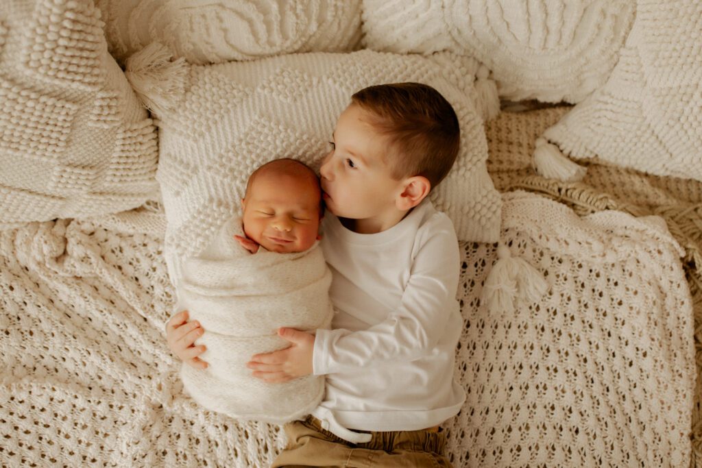 Toddler boy snuggling his new baby brother and kissing the top of his head