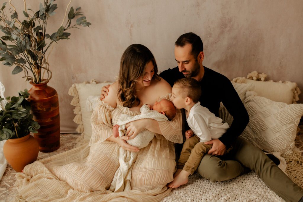 Mom and Dad in cozy lifestyle studio setup hold their newborn while their toddler son kisses the top of baby's head