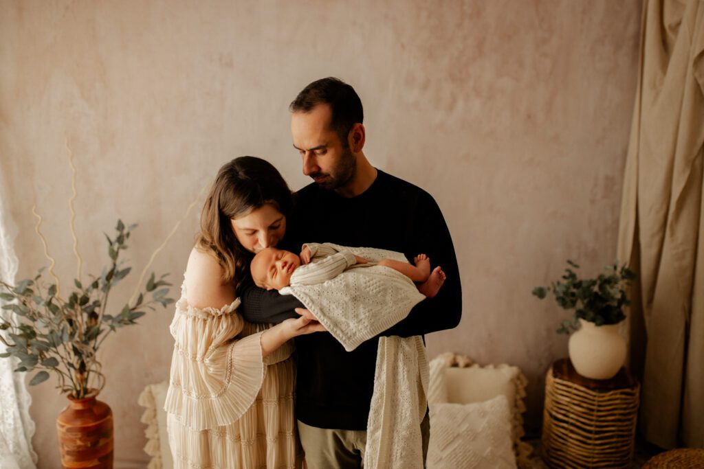 Dad standing in cozy alcove holding his newborn son while Mom stands next to him and leans down to kiss baby's head
